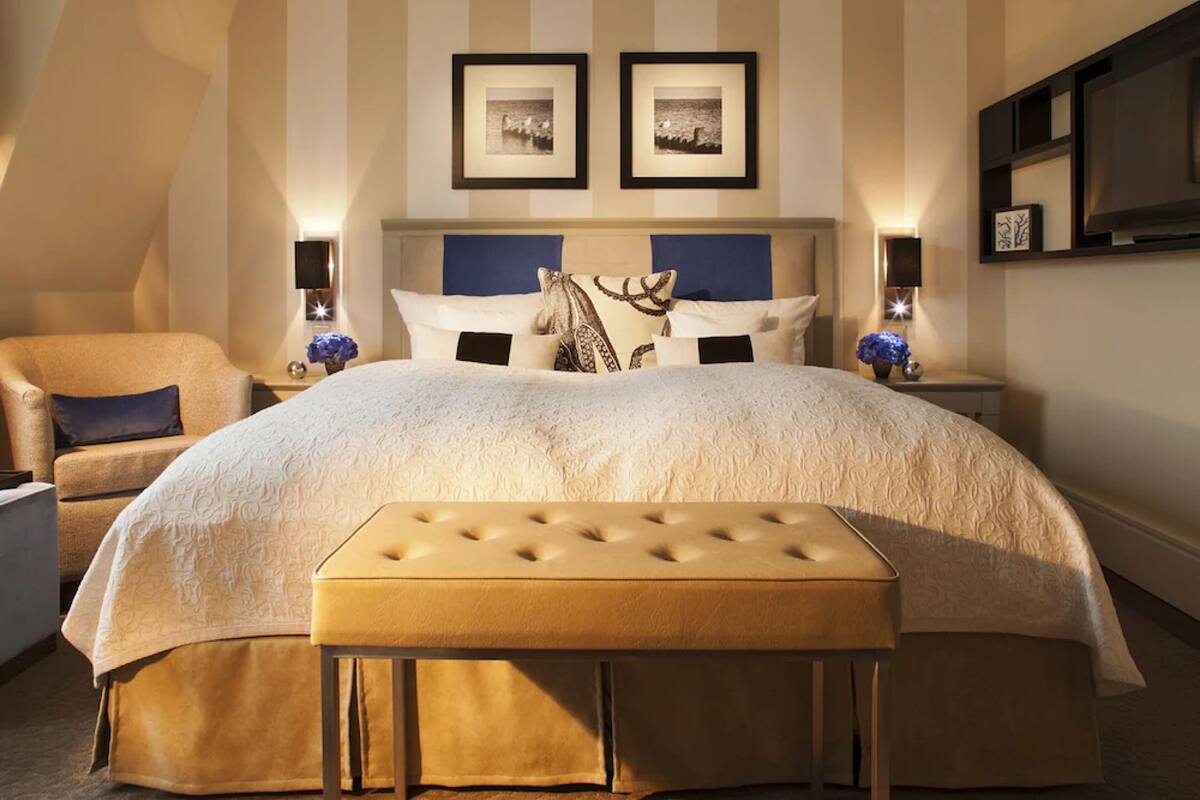 Una cama grande con cabecera capitonada, un banco y una silla en una habitación con paredes a rayas y cuadros enmarcados sobre la cama.