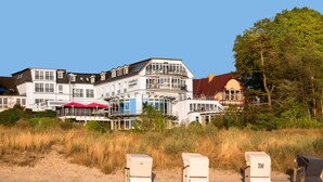 A beachfront hotel with multiple buildings, including a prominent structure with a red roof and large windows. There are beach chairs and umbrellas visible in the foreground. - Strandhotel Ostseeblick (Heringsdorf)