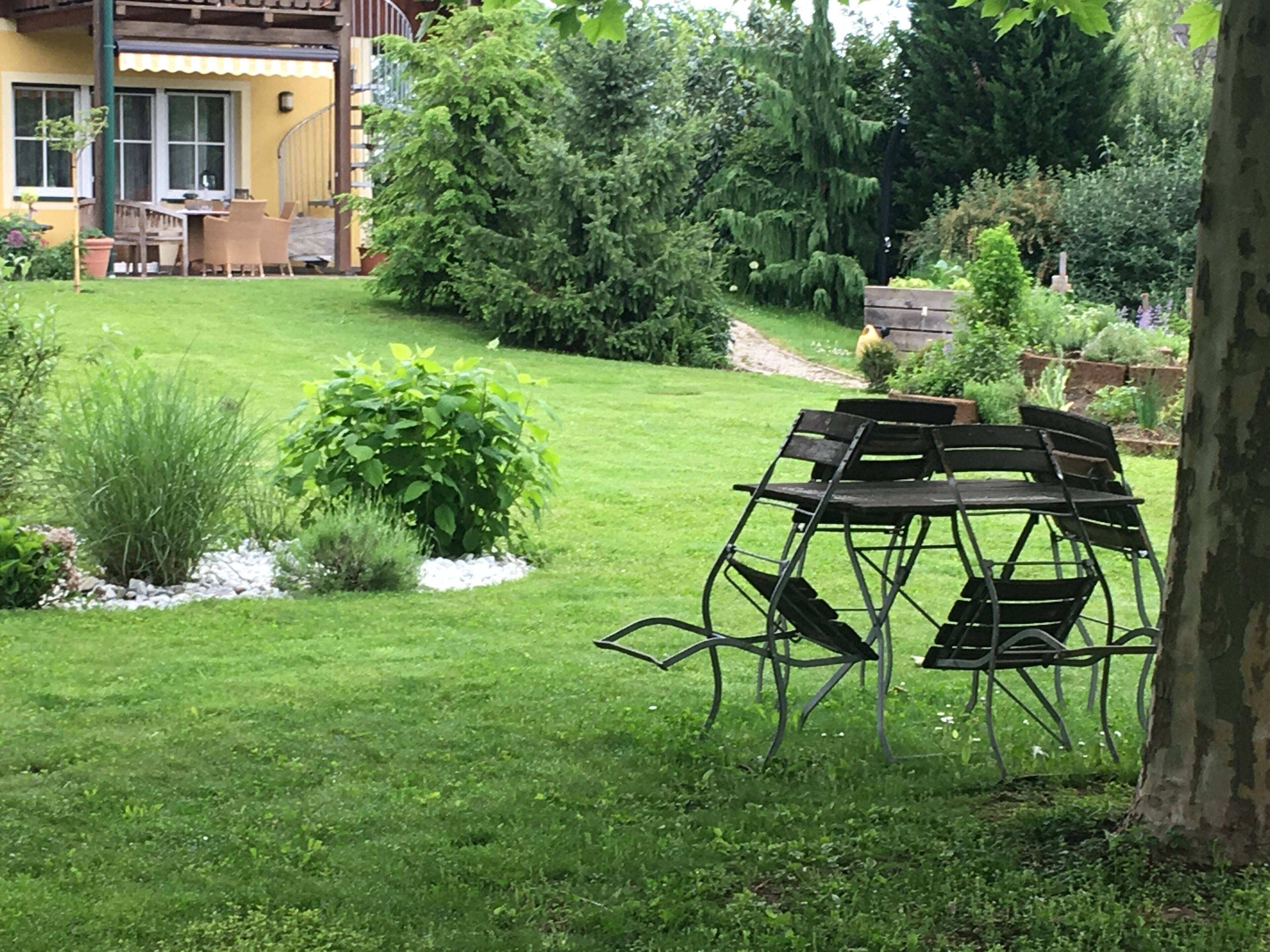 A garden with a wooden table and chairs, a yellow building, and various plants.