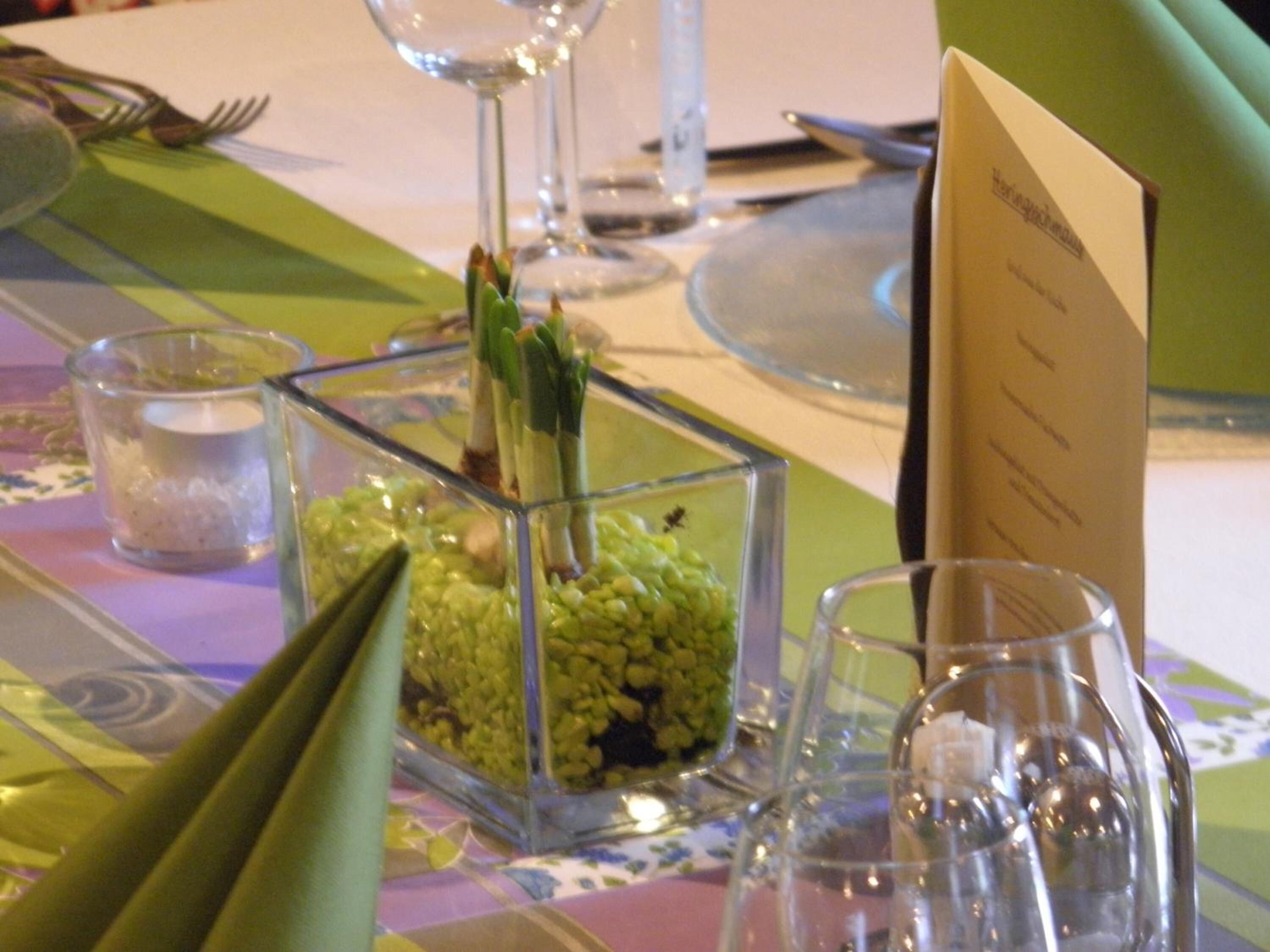 A table setting with a clear glass centerpiece containing green vegetables, a menu card, and various tableware.