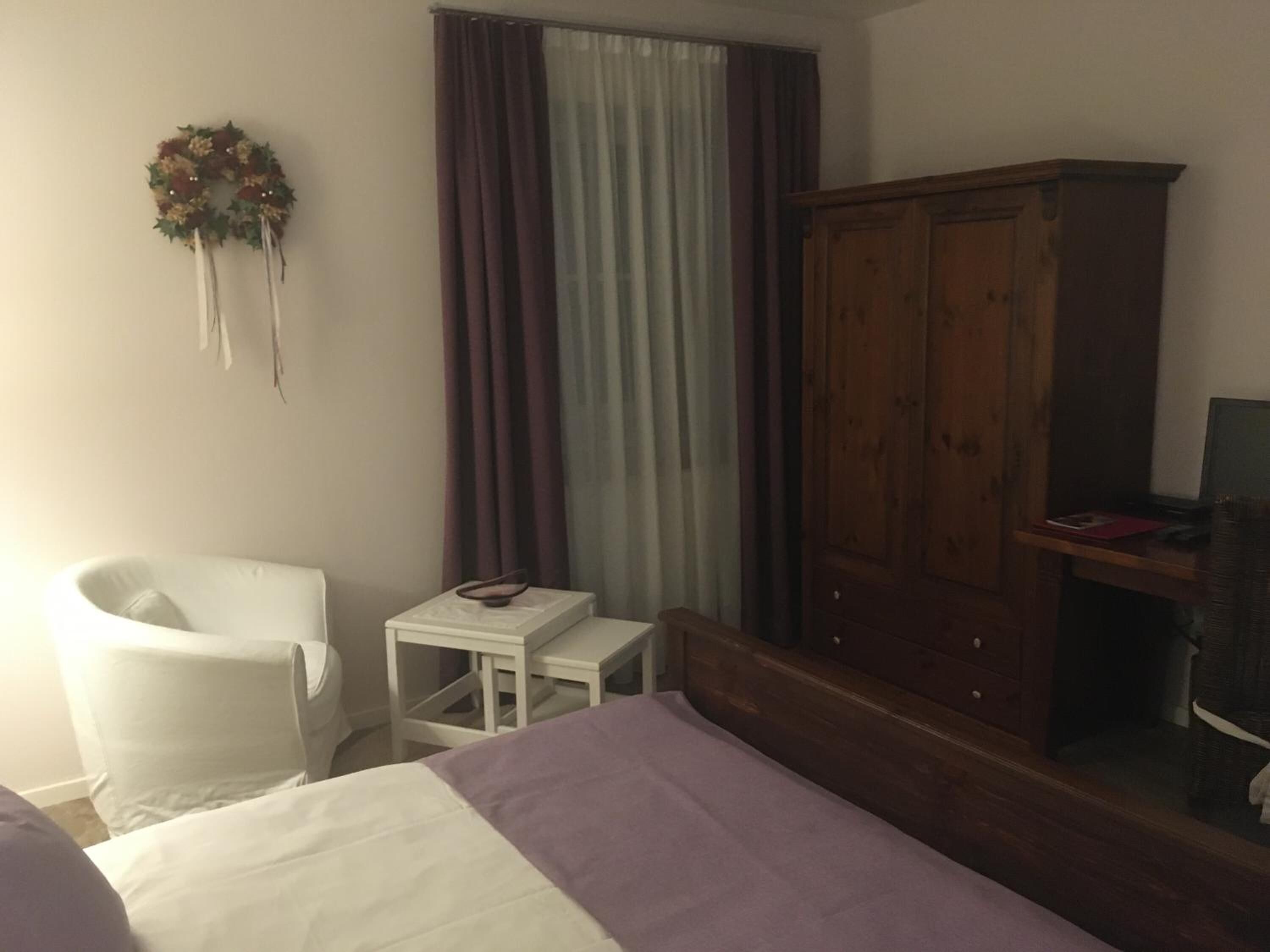 A bedroom with a bed, a white chair, a small table, a wooden wardrobe, and a television on a stand.