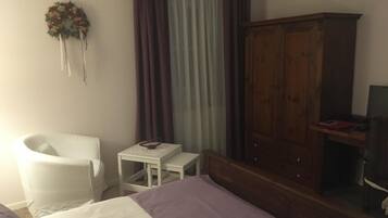 A bedroom with a bed, a white chair, a small table, a wooden wardrobe, and a television on a stand.