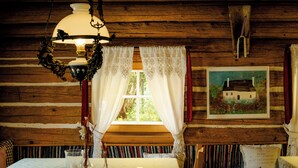 Ein rustikales Holzhaus-Interieur mit Esstisch, einer Hängelampe, Spitzenvorhängen und einem Bilderrahmen mit einem Hausgemälde.