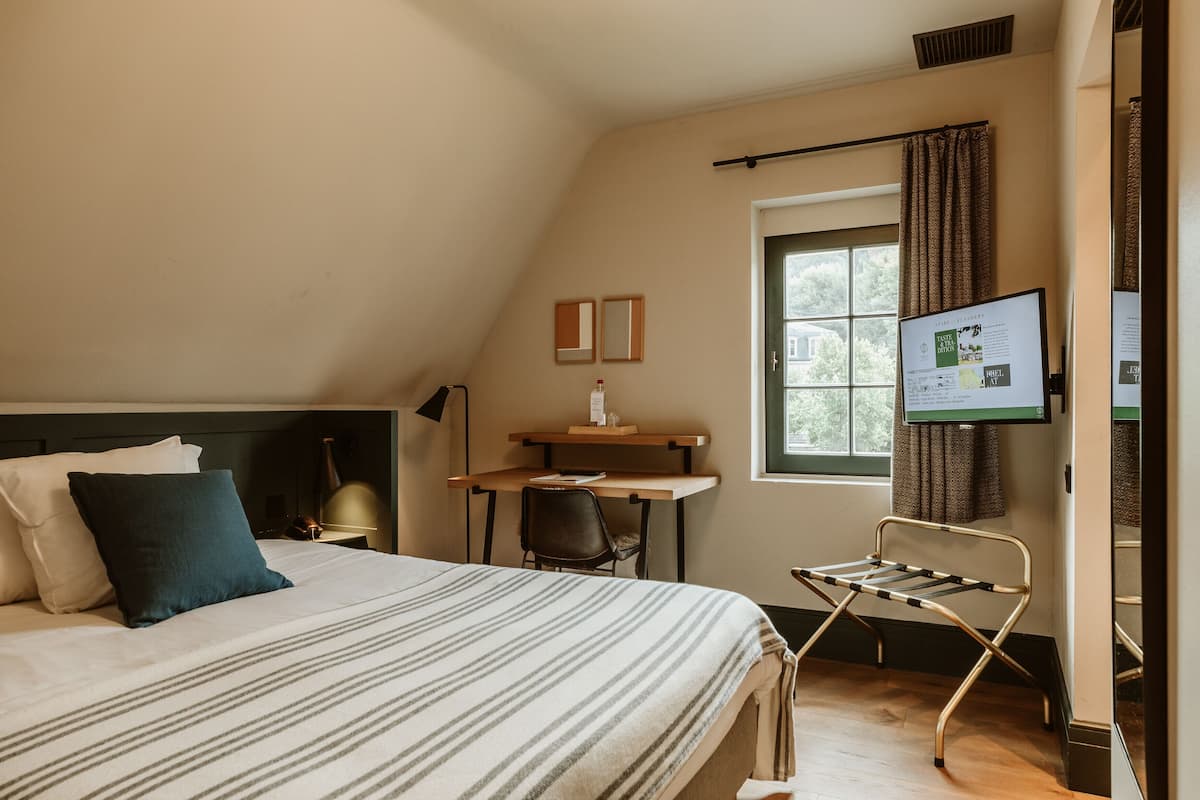 A bedroom with a bed, a desk, and a chair. There is a window with curtains and a TV mounted on the wall.