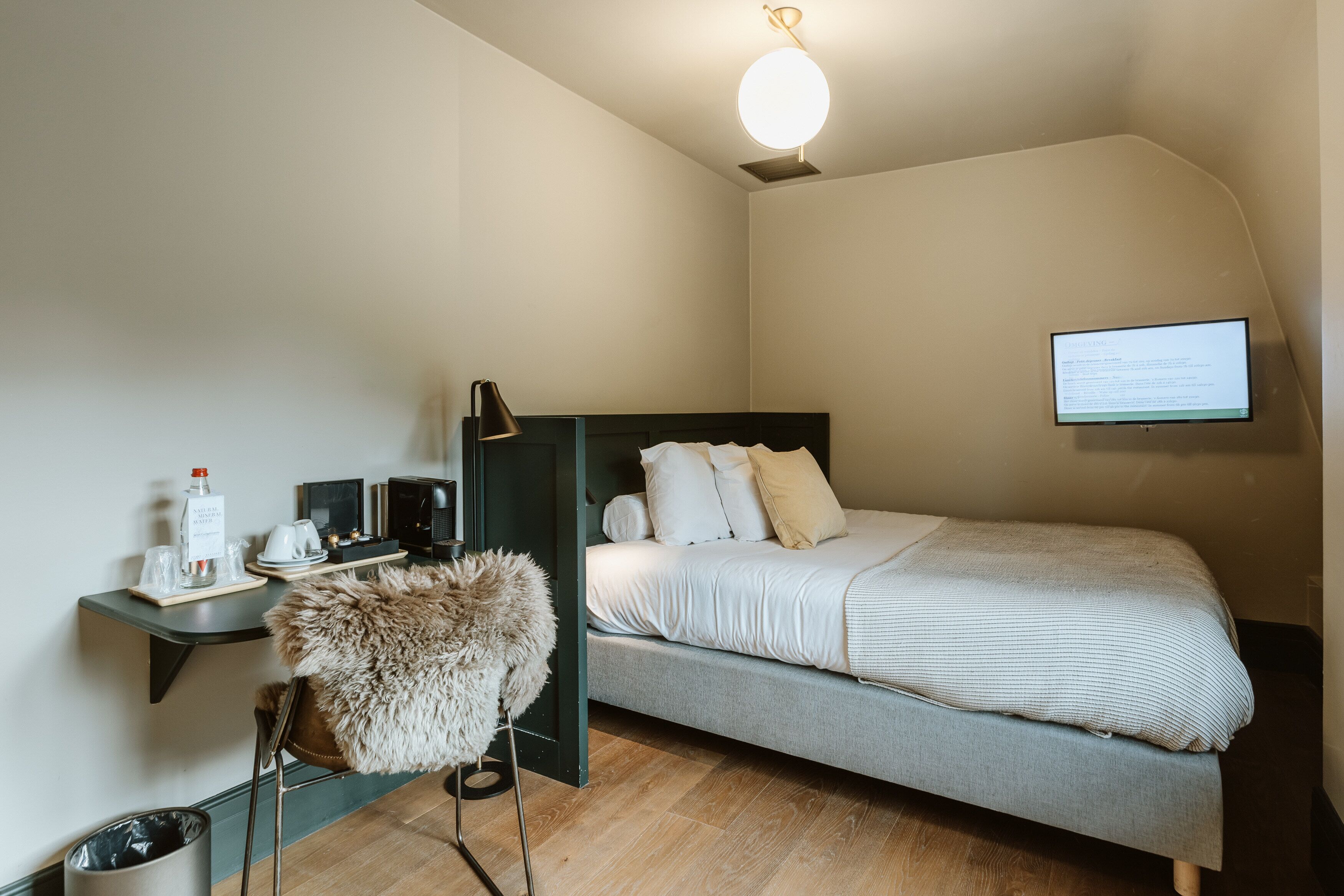 Ett kompakt hotellrum med en säng, ett skrivbord med en dator, en stol, ett litet bord med en flaska och glas, och en tv på väggen.
