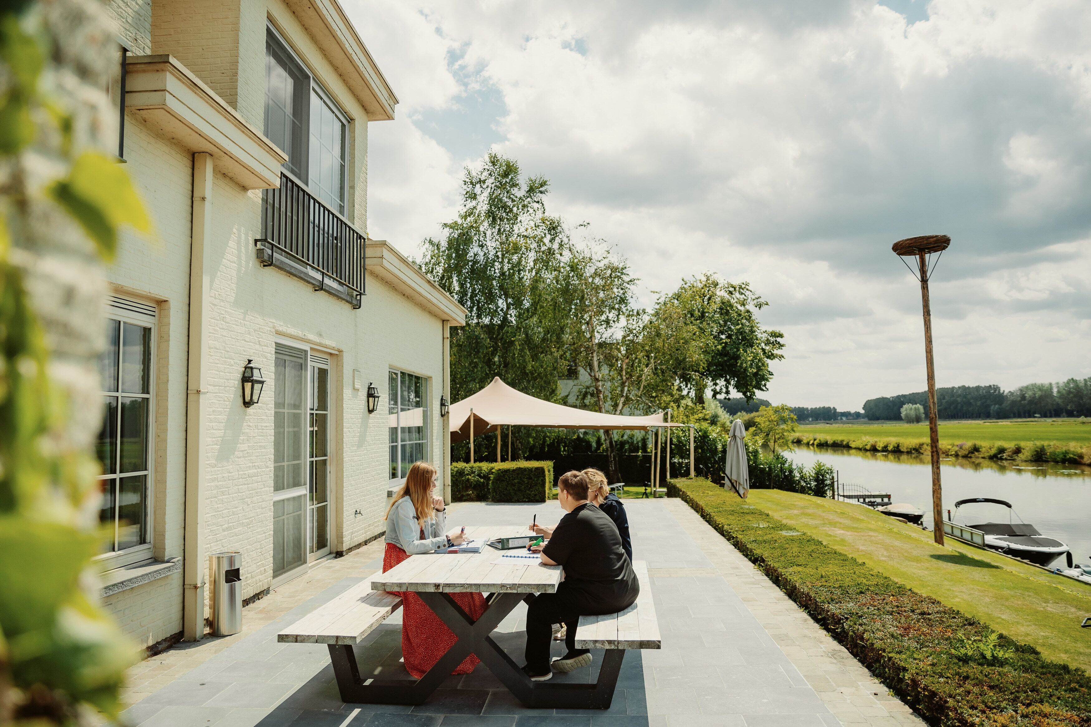 Une terrasse avec une table où sont assis deux personnes, un bâtiment blanc doté de grandes fenêtres et une rivière en arrière-plan.