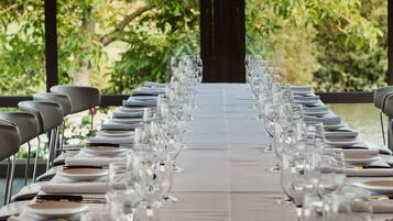 A long dining table set with multiple glasses and plates, surrounded by chairs, with a view of greenery and a structure in the background.