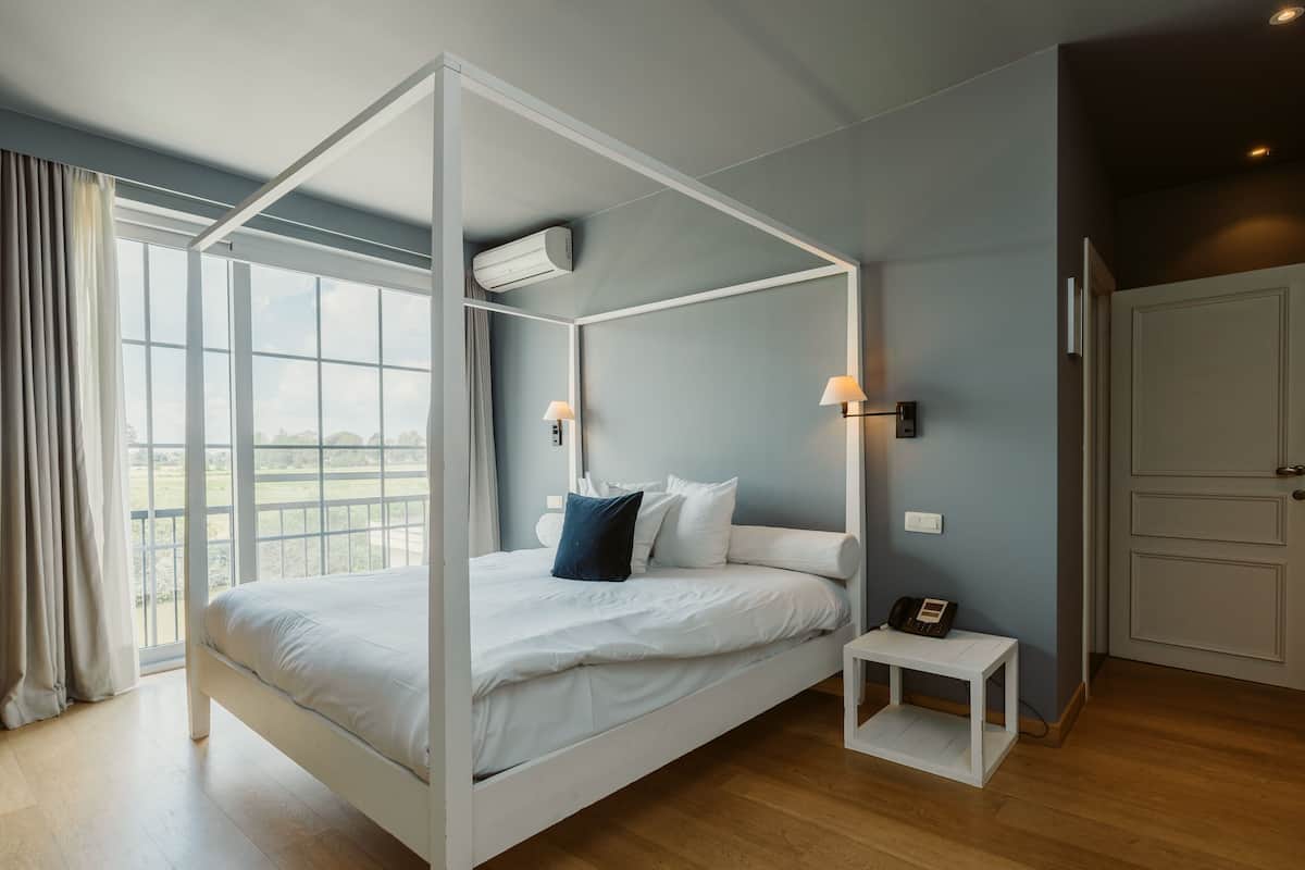 A modern bedroom with a four-poster bed, a bedside table with a phone, and a view of the outdoors through large windows.