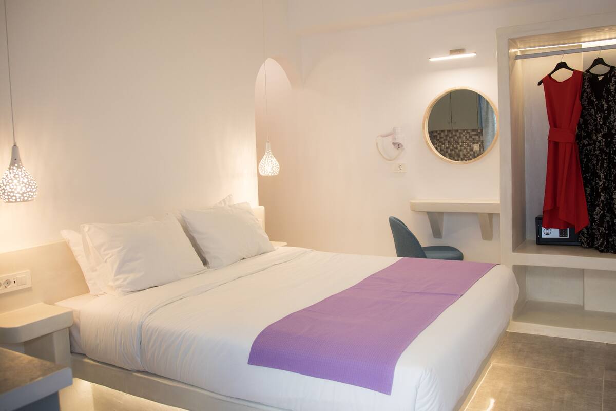 A hotel room with a bed, a desk, a chair, and a wardrobe with clothes hanging.