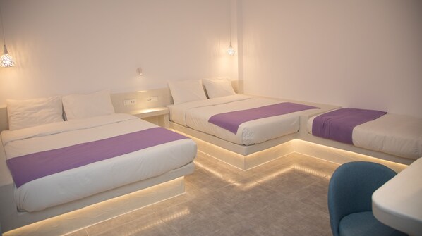 A hotel room with three beds arranged in a U-shape, each with white bedding and purple accents. There are wall-mounted lights and a visible power outlet on the nightstand.