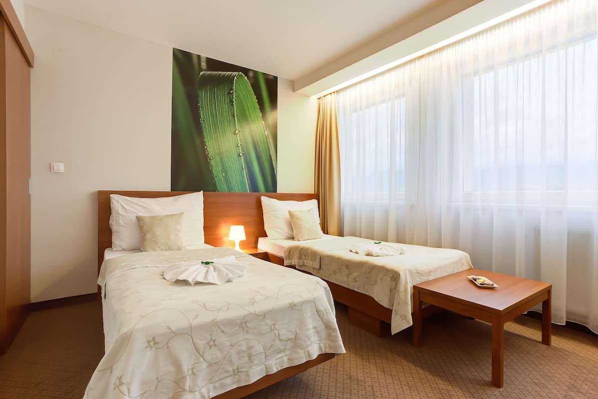 Habitación de hotel con dos camas, una mesita de noche de madera, una mesa, una lámpara y un ventanal grande con cortinas.