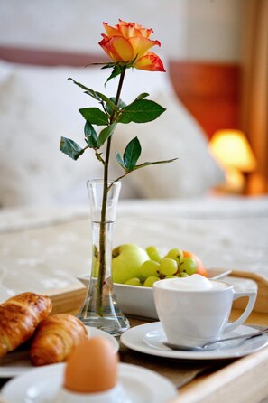 Bandeja de desayuno con cruasanes, una taza de café, un bol de fruta y una florera con una rosa.