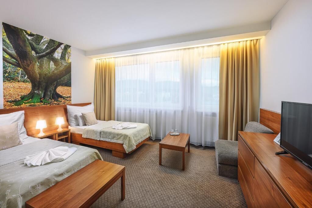 A hotel room with two beds, a TV, a desk, and a large window with curtains.