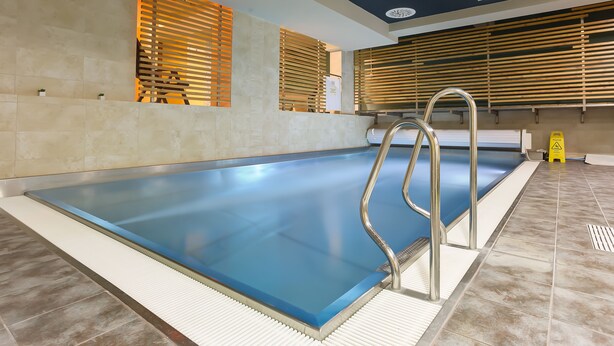 Una piscina con interior azul, rodeada de baldosas color beige y listones de madera en las paredes.