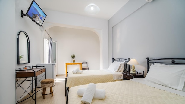 A hotel room with two beds, a desk, a chair, a mirror, a TV, and a small table with a plant. - Emmanouela Studios (Santorini)
