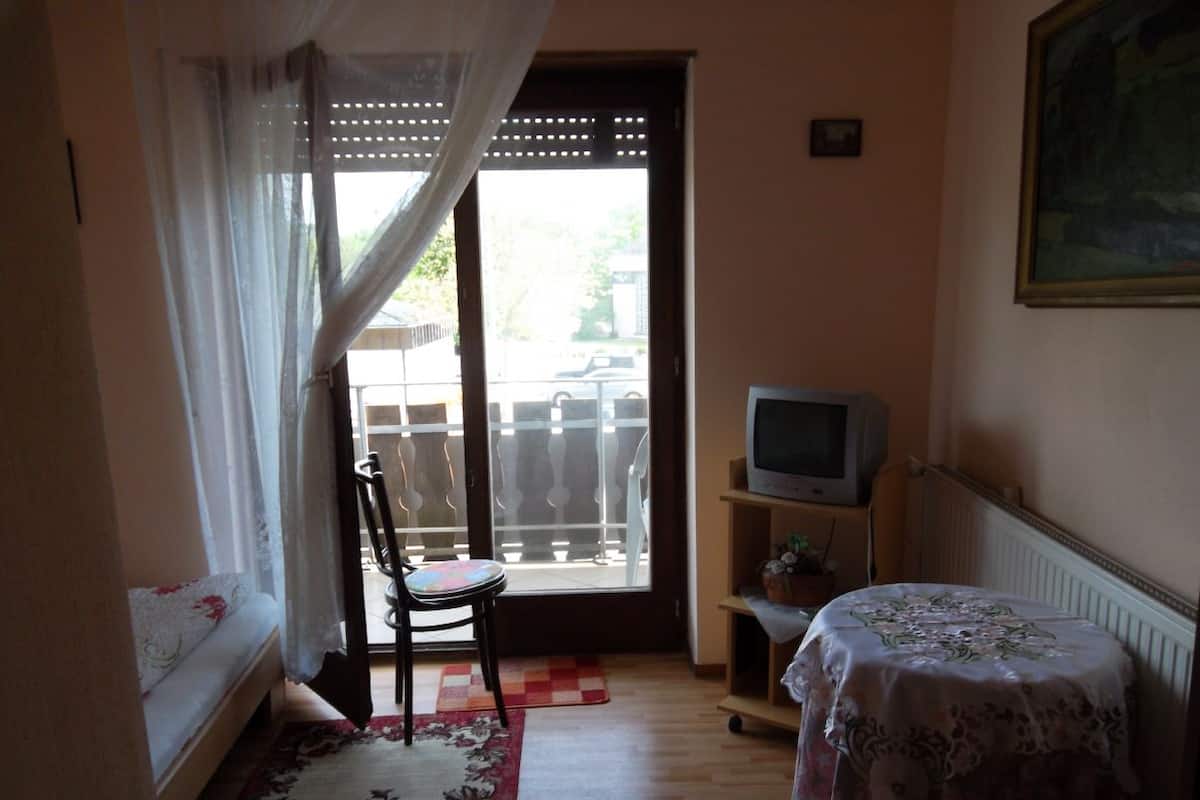 Une petite pièce comprenant un lit, une chaise, un téléviseur et une fenêtre avec des rideaux.