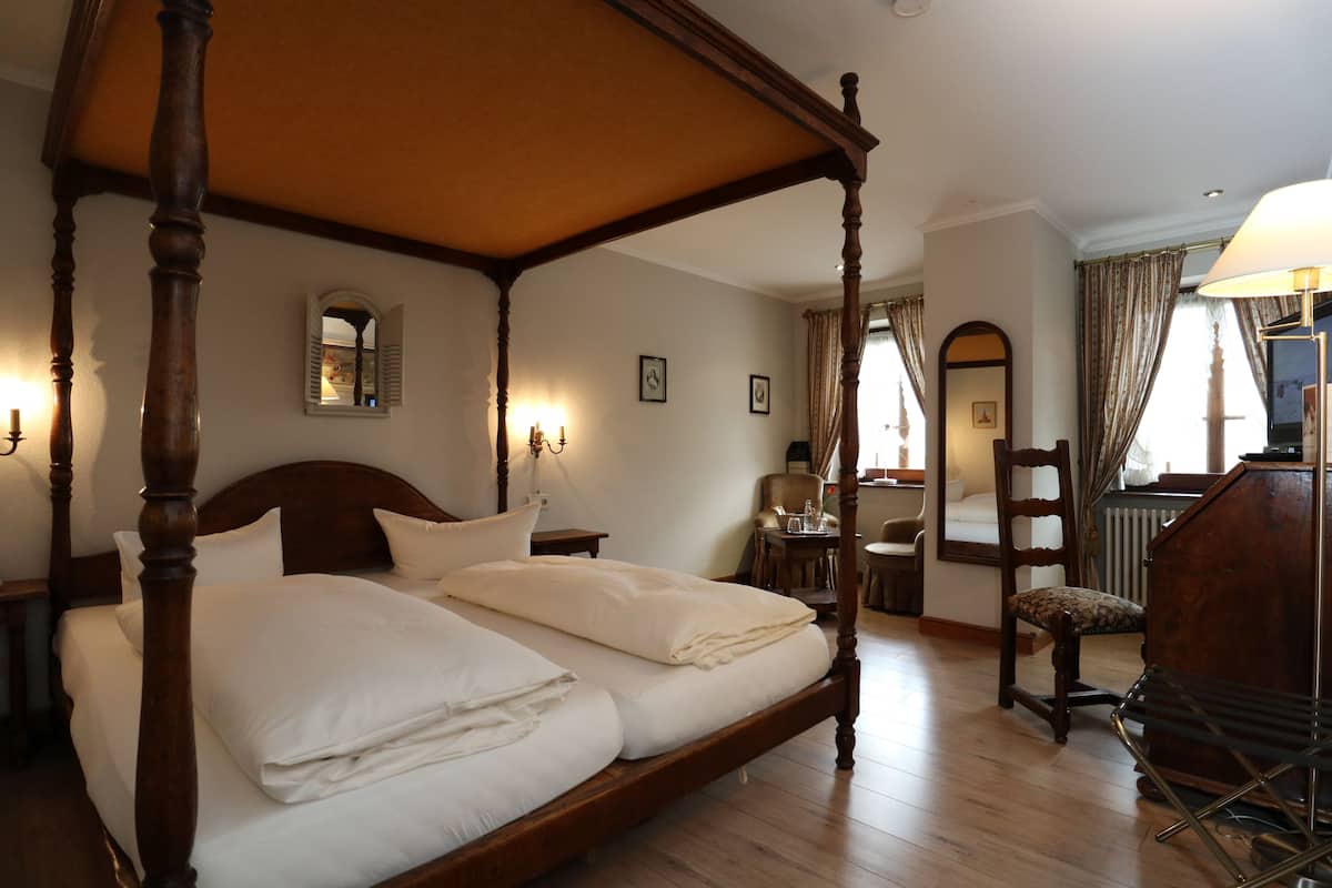 A four-poster bed with a wooden frame and white linens, a wooden chair, a small table with a lamp, and a mirror on the wall.