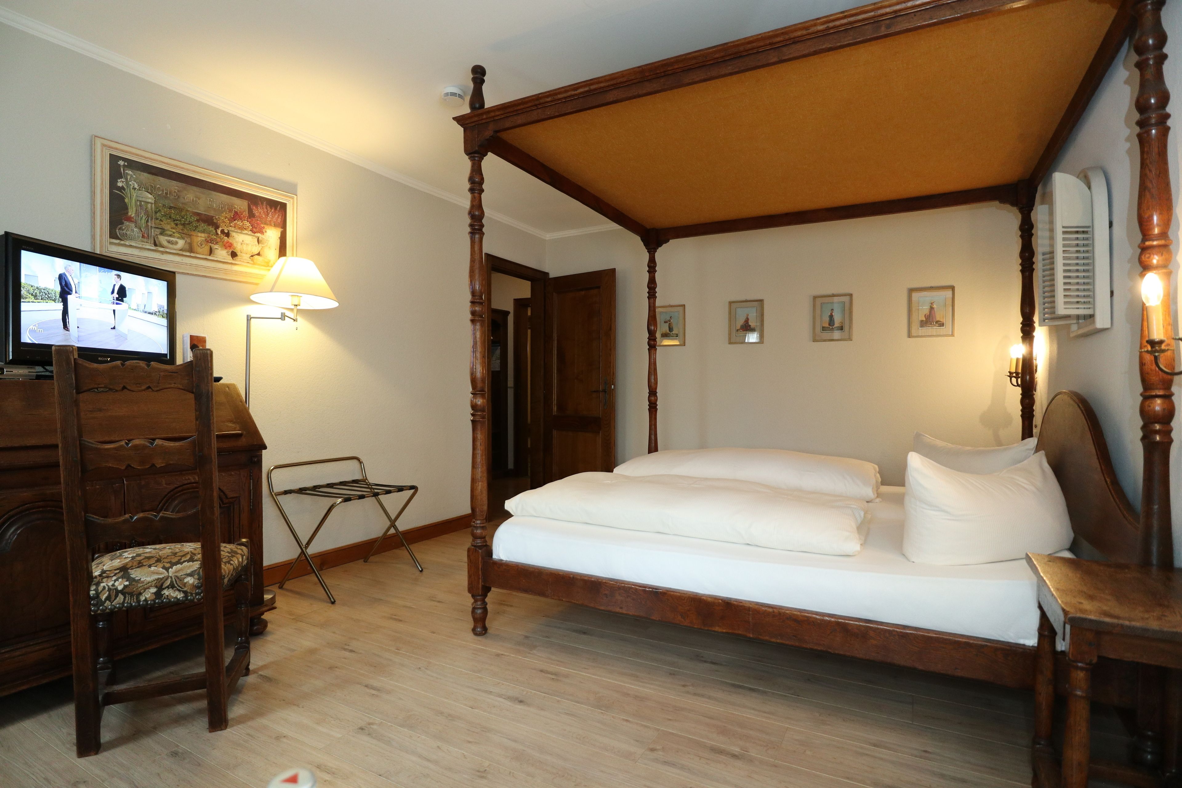 Una cama con dosel y estructura de madera, un televisor sobre un soporte de madera, una silla plegable y una lámpara fijada a la pared.