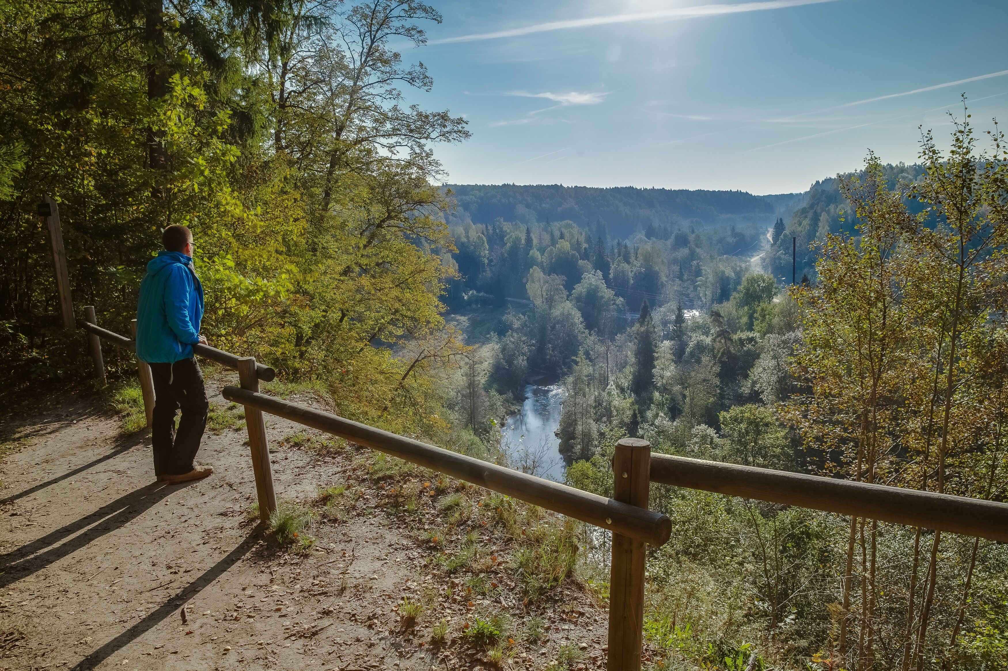 A person standing on a dirt path with a wooden railing, overlooking a valley with a river and dense forest.