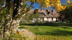 A traditional building with a sloped roof, surrounded by a grassy area with scattered fallen leaves. - Karlamuiza Country Hotel (Cesis Municipality)