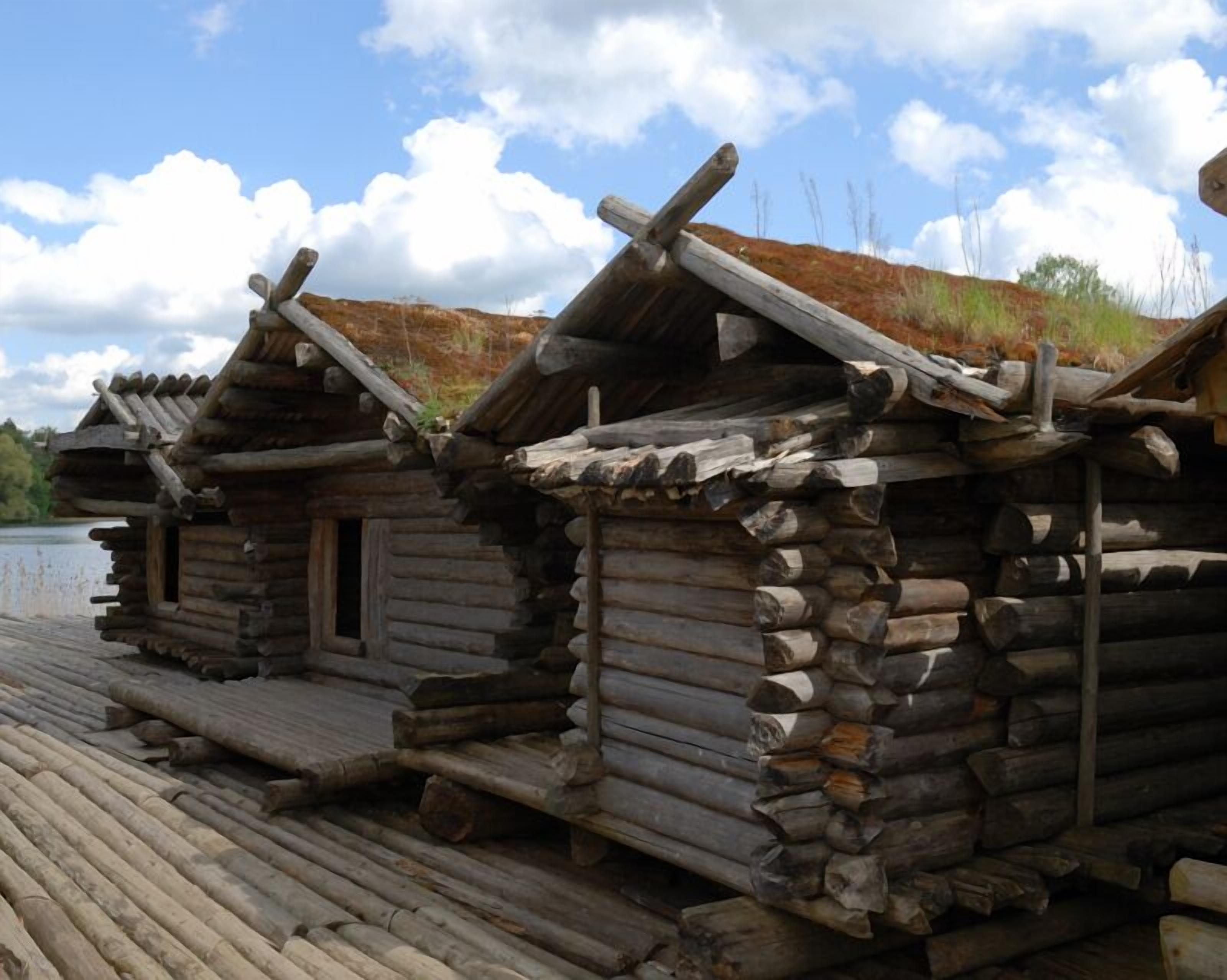 A row of log cabins with a wooden deck in front, set against a backdrop of a clear sky and distant trees.