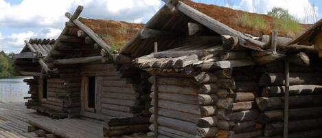 A row of log cabins with a wooden deck in front, set against a backdrop of a clear sky and distant trees.