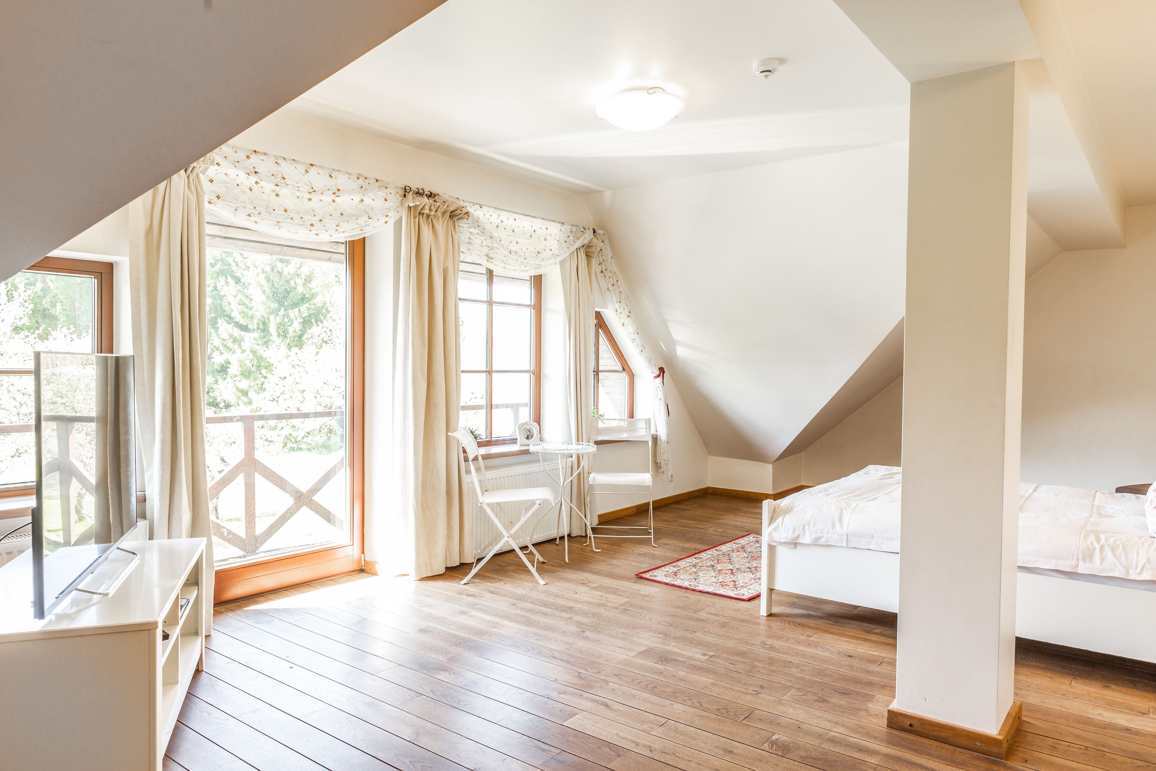 Ein Zimmer mit schrägem Dach, einem Bett, einem Schreibtisch mit Stuhl und einem Fenster mit Vorhängen.