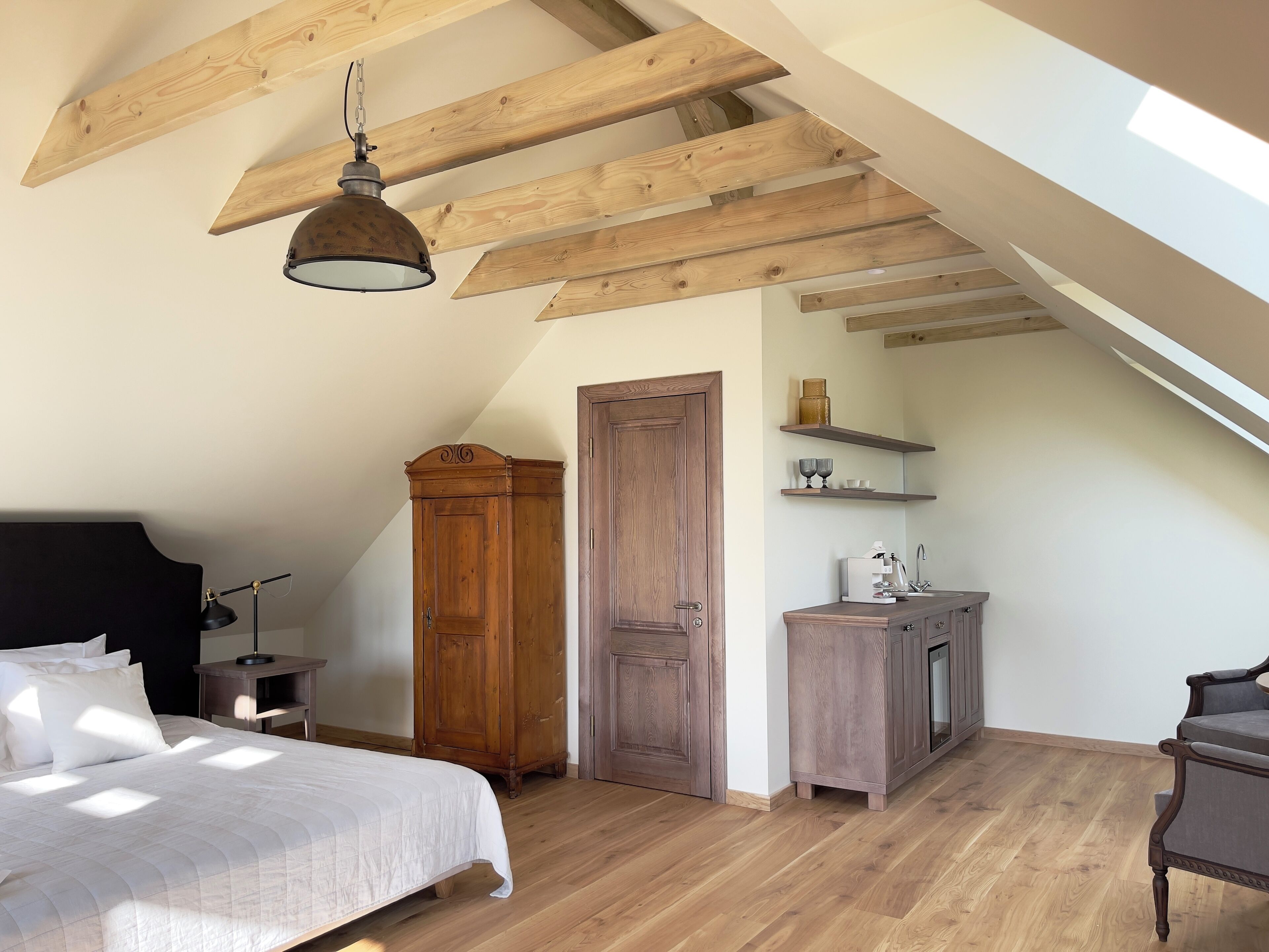 A bedroom with a sloped ceiling, wooden beams, a bed with white bedding, a wardrobe, a door, a small kitchenette, and a chair.