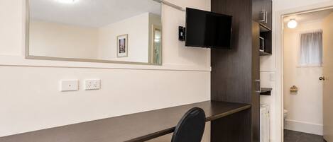 A modern hotel room with a long desk, a chair, a flat-screen TV, a large mirror, and a small kitchenette.