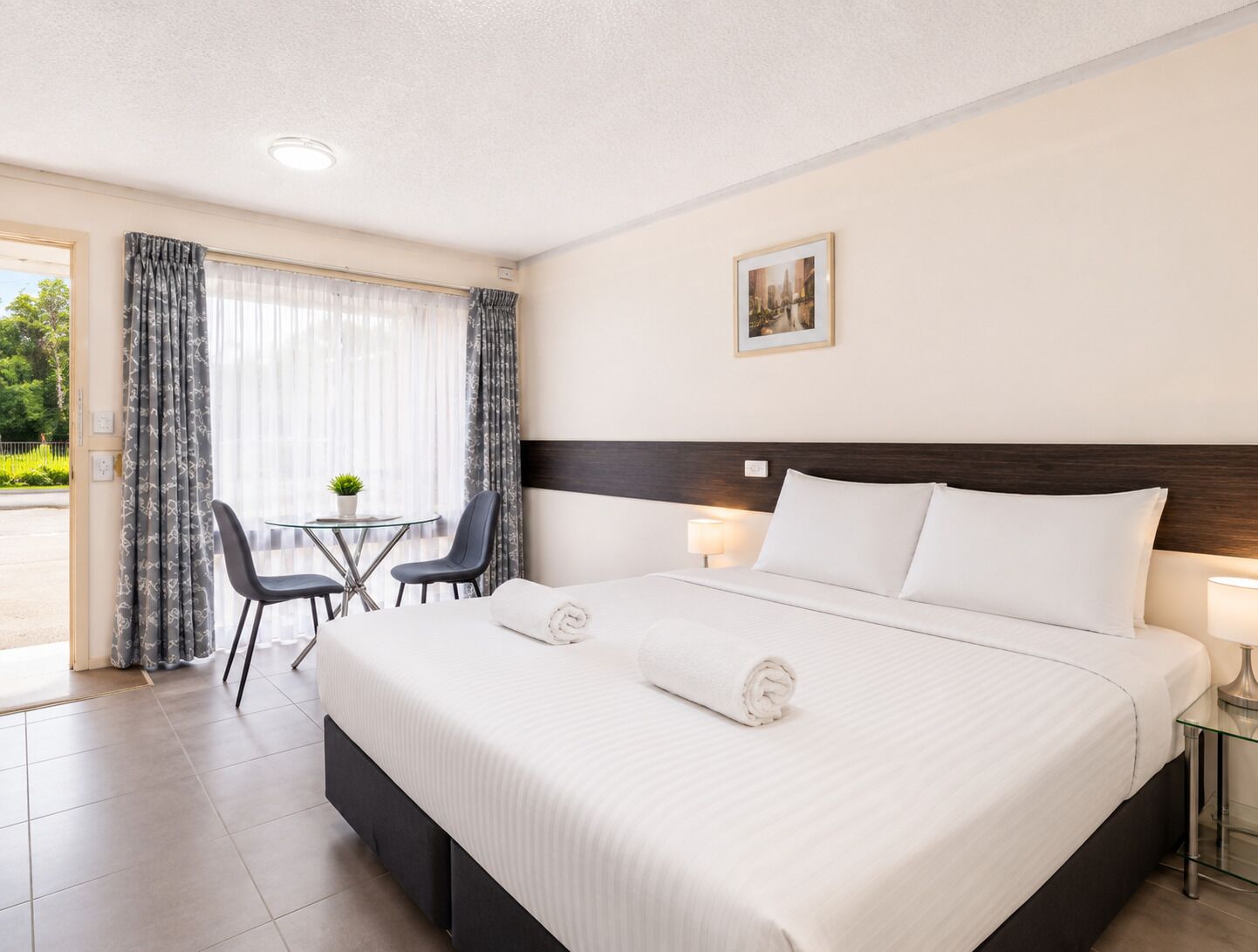 Quarto de hotel com uma cama grande, duas luminárias de cabeceira, uma mesinha com cadeiras e uma janela com cortinas.