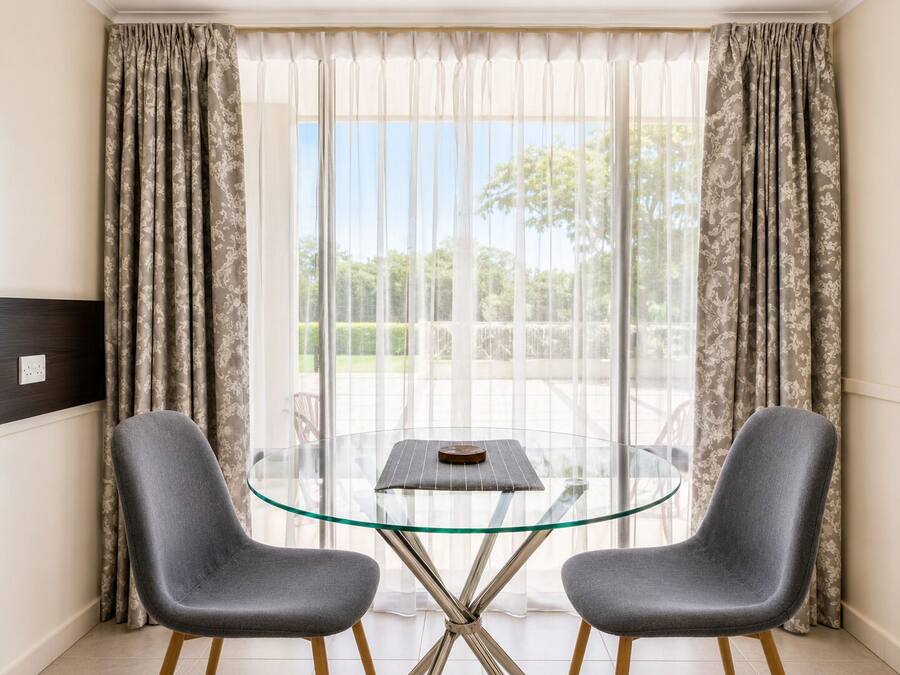 A room with a glass table and two chairs, a window with curtains, and a view of greenery outside.