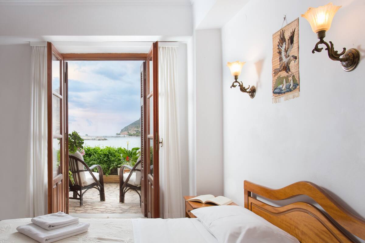 A hotel room with a bed, a nightstand, a reading lamp, and a view of the sea through an open door.