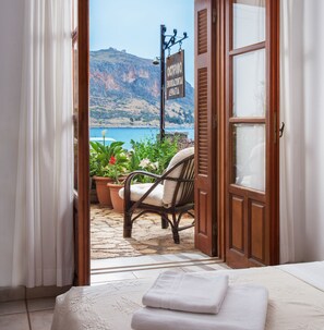 A room with a view of a coastal landscape, a wicker chair, and a sign indicating a restaurant. - Petrino Guesthouse (Monemvasia)