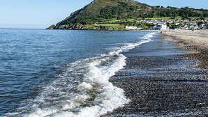 Uma praia de seixos com ondas suaves a beijar a costa, uma colina distante coberta de vegetação e uma cidade ao fundo.