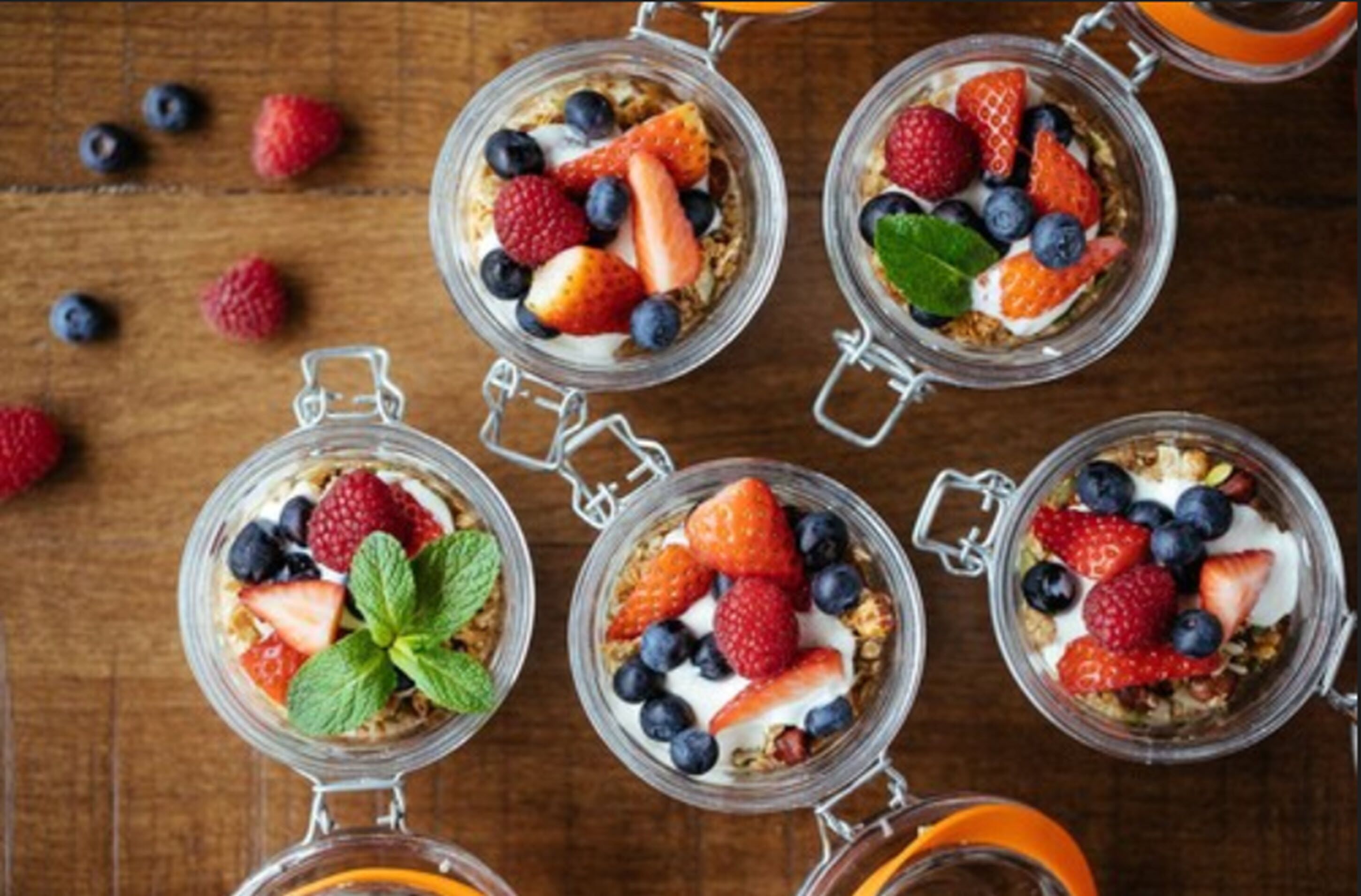 Five glass jars with lids, each containing a layered dessert with strawberries, blueberries, and a mint leaf.