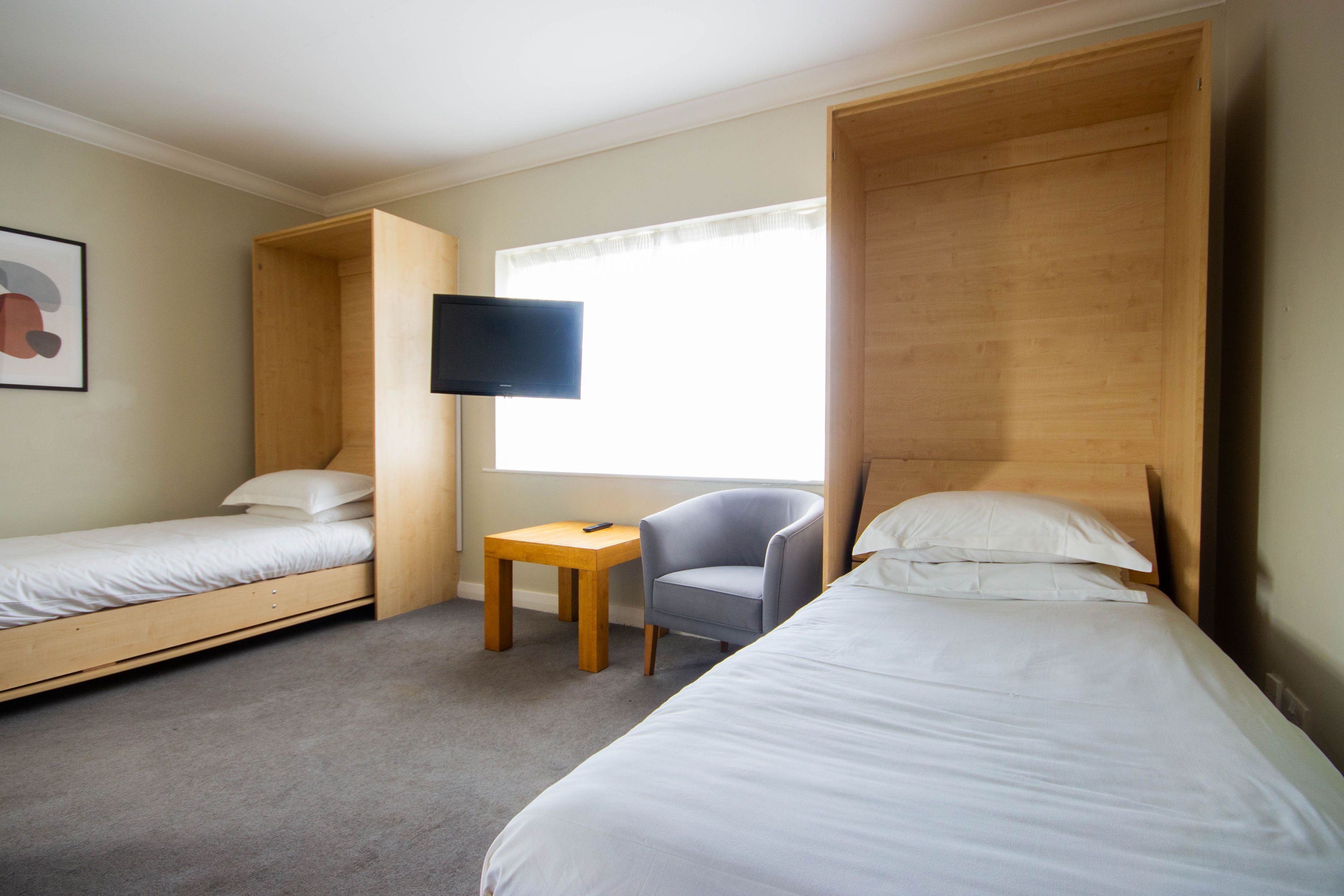 A hotel room with two single beds, a small table, a chair, a television, and a window with curtains.
