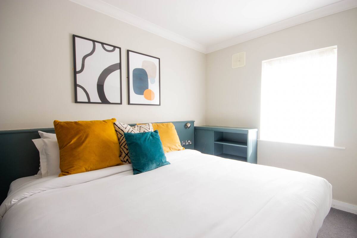 A neatly made bed with white bedding and colorful pillows, a teal nightstand, and abstract wall art.