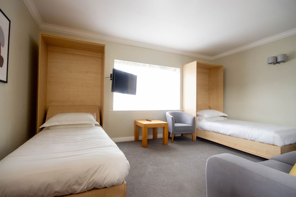 A room with two single beds, a small table, a chair, and a television mounted on the wall.