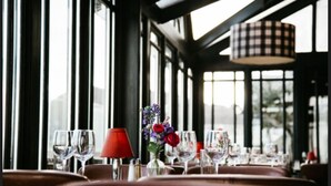 Interior de restaurante com grandes janelas, mesa posta com taças de vinho, abajur vermelho e um vaso com flores.