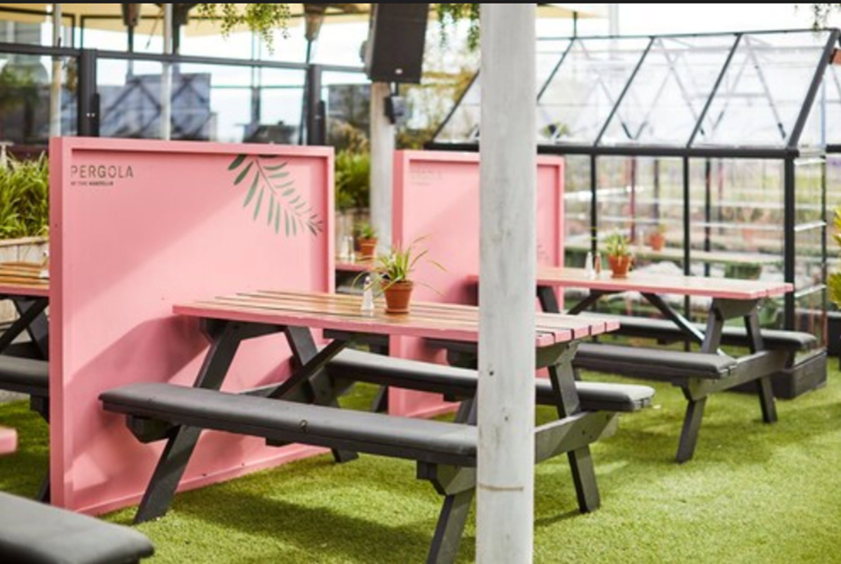 An outdoor seating area with pink tables and benches, surrounded by greenery and a greenhouse structure.