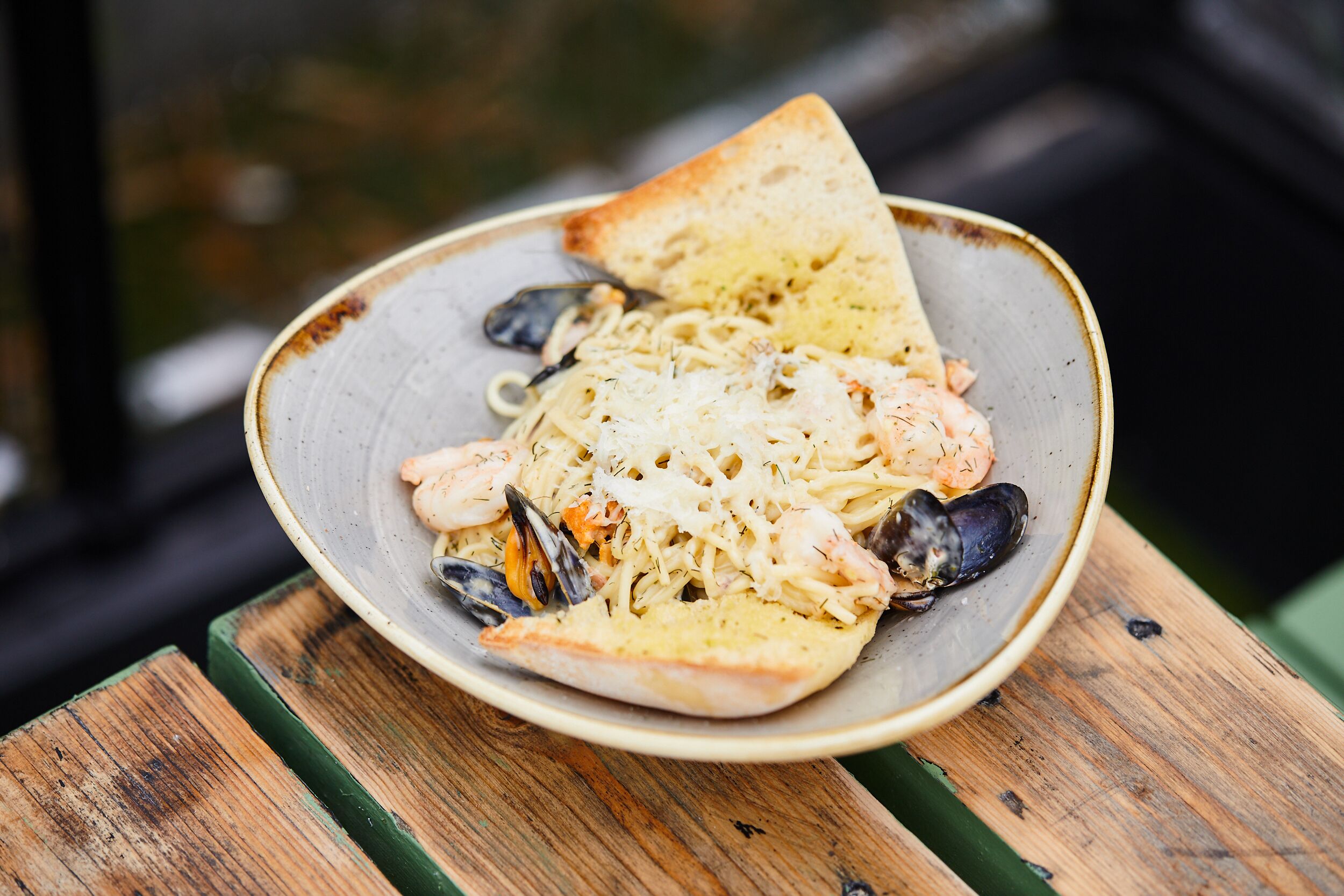 Un plato de pasta con mariscos, con mejillones y camarones, servido en un plato de cerámica y acompañado de una rebanada de pan sobre una mesa de madera.