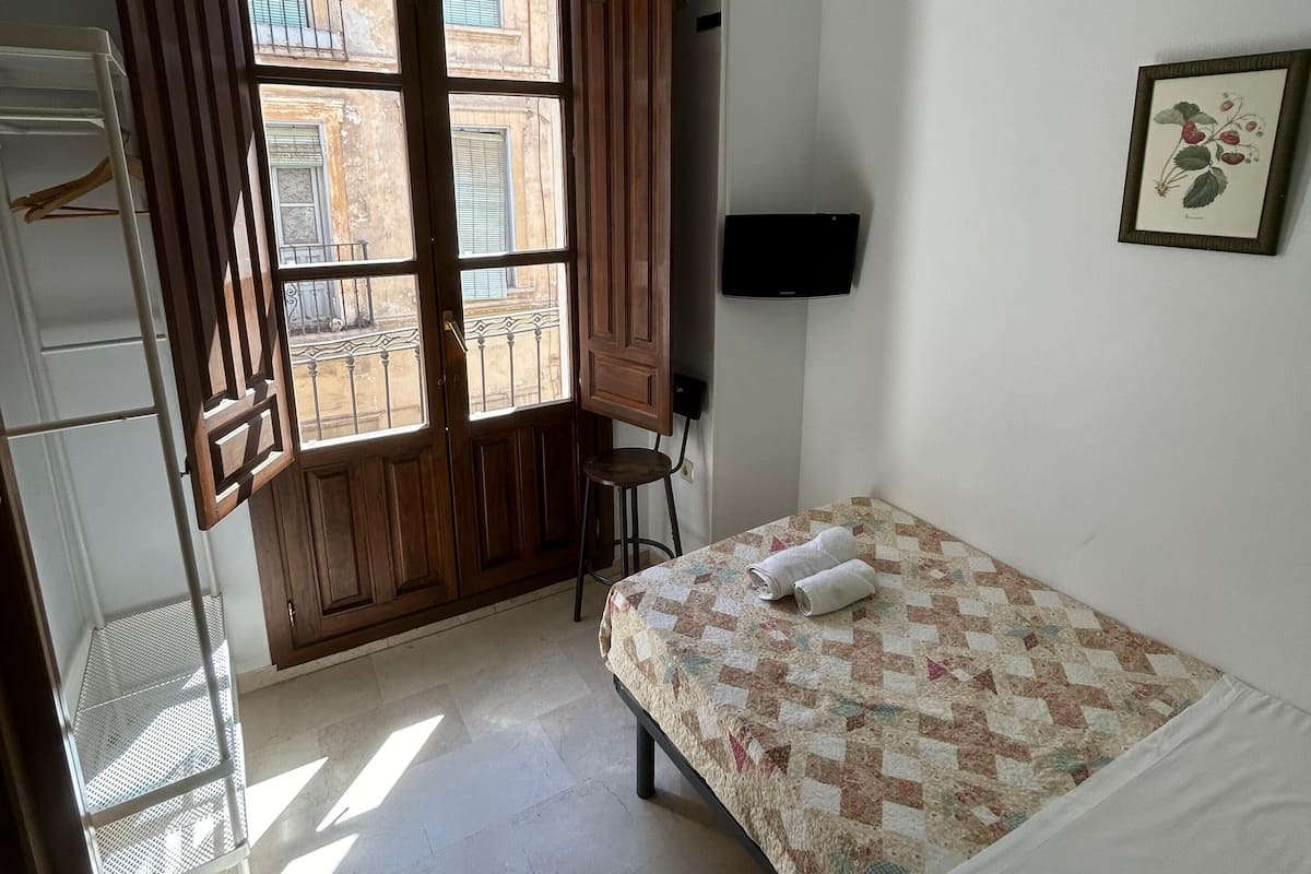 A bedroom with a bed, a TV, a window with wooden shutters, and a balcony.