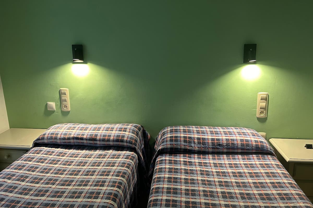 Twin beds with plaid bedding, two bedside tables, and wall-mounted light fixtures in a room with green walls.