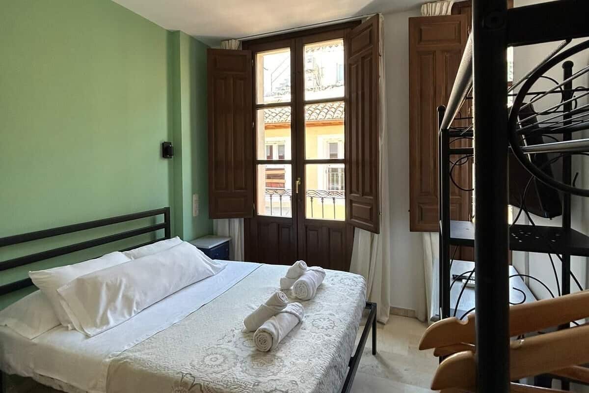 A room with a bed, a window with wooden shutters, a ceiling fan, and a spiral staircase.