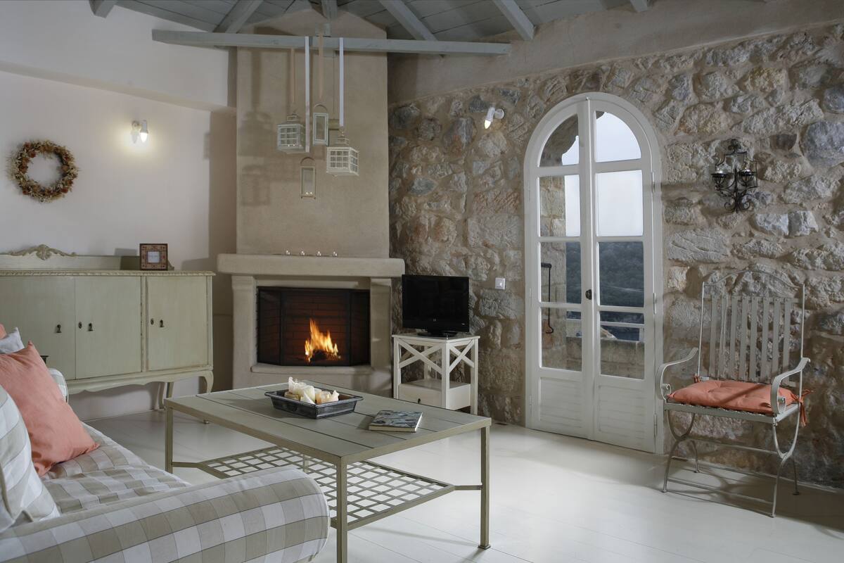 A cozy living room with a fireplace, a television, a stone wall, and a view through an arched doorway.