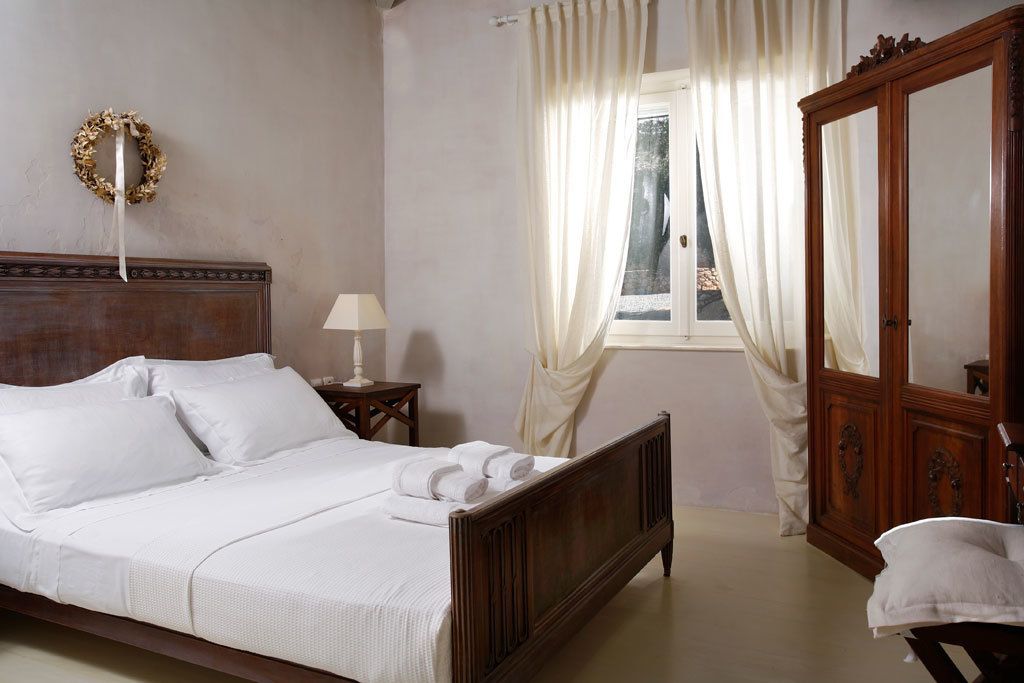 Un lit bien fait, avec du linge de lit blanc, une tête de lit en bois, une table de chevet avec une lampe, un grand miroir et une fenêtre ornée de rideaux légers.