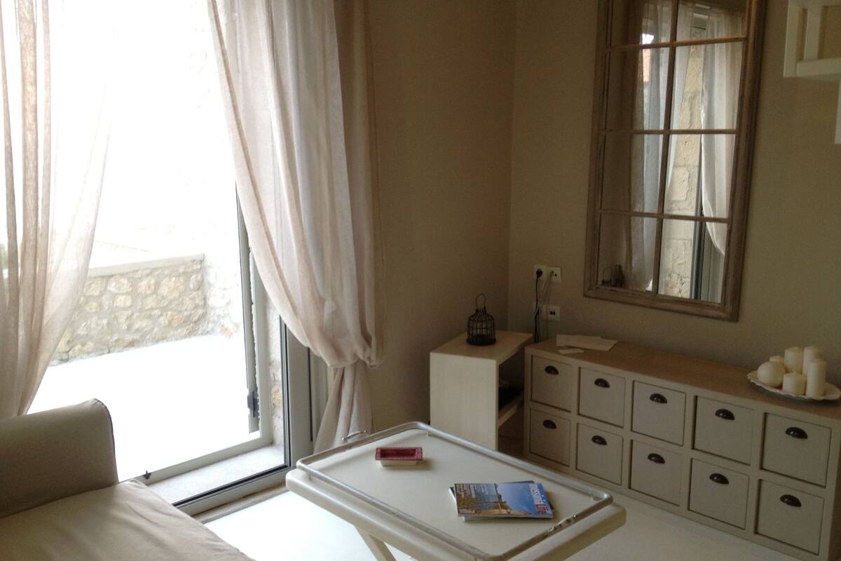 A bedroom with a bed, a bedside table with a book, a dresser with drawers, a mirror, and a window with curtains.