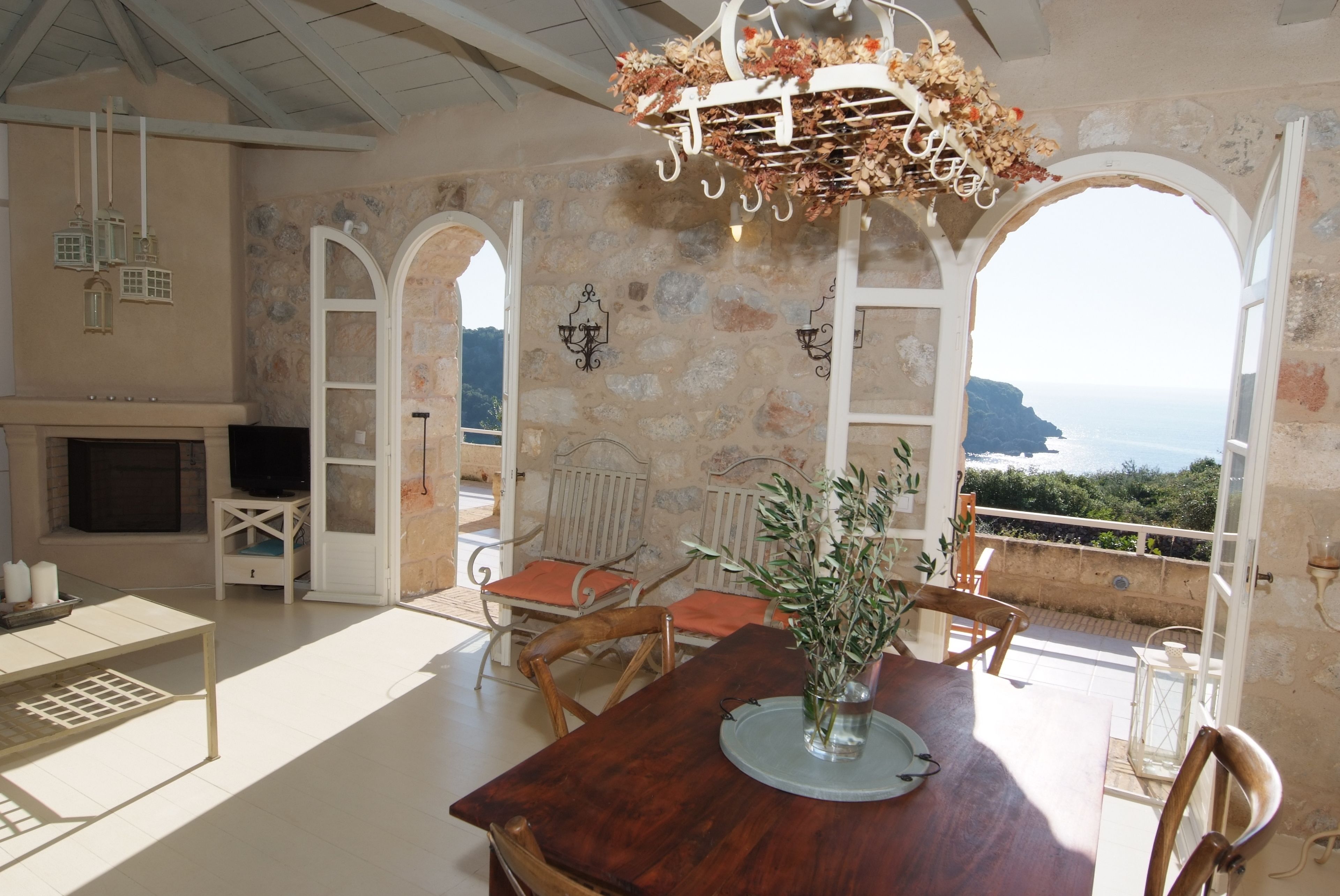 Uma sala de jantar com mesa de madeira, cadeiras, um vaso com flores e vista para o mar através de uma abertura.
