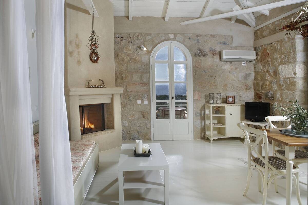 A room with a stone wall, a fireplace, a television, and a dining area with white furniture.