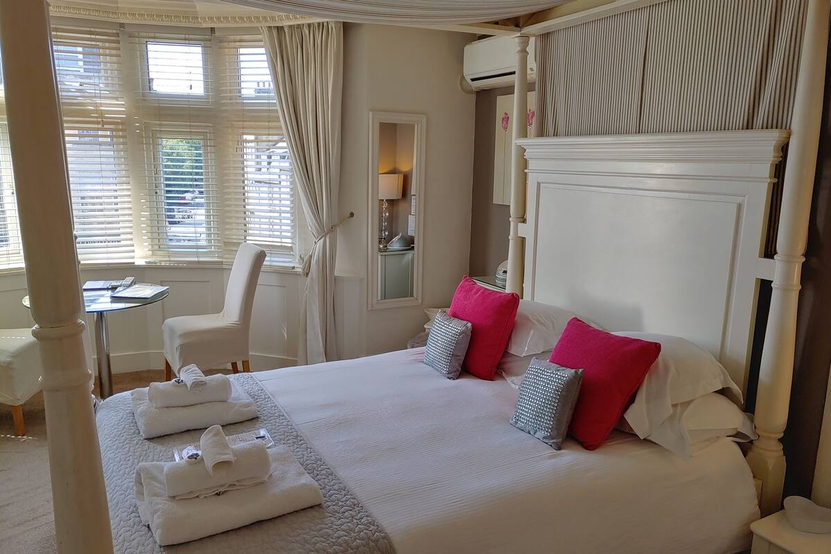 A neatly made bed with white linens and multiple pillows, a small table with a lamp, and a chair in a well-lit room with large windows and curtains.